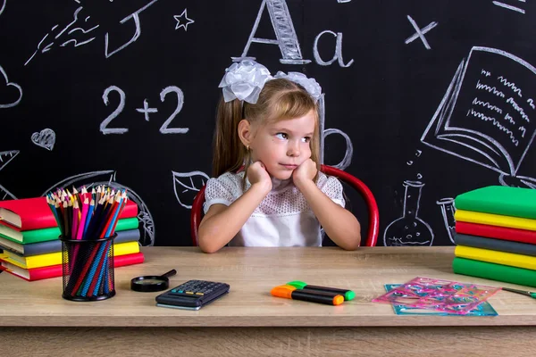 Colegiala aburrida sentada en el escritorio con libros, útiles escolares, sosteniendo sus dos manos sobre las mejillas, mirando a un lado — Foto de Stock