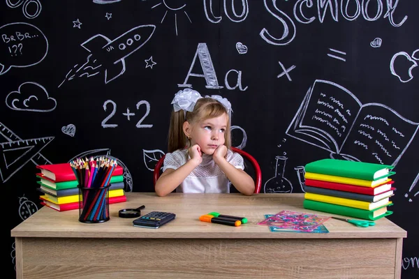 Escolar aburrida y soñadora sentada en el escritorio con libros, útiles escolares, sosteniendo sus dos manos sobre las mejillas, mirando hacia arriba — Foto de Stock