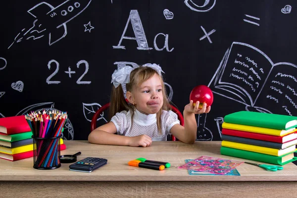 Colegiala hambrienta sentada en el escritorio con libros, útiles escolares, sosteniendo en su brazo manzana roja lista para morder — Foto de Stock