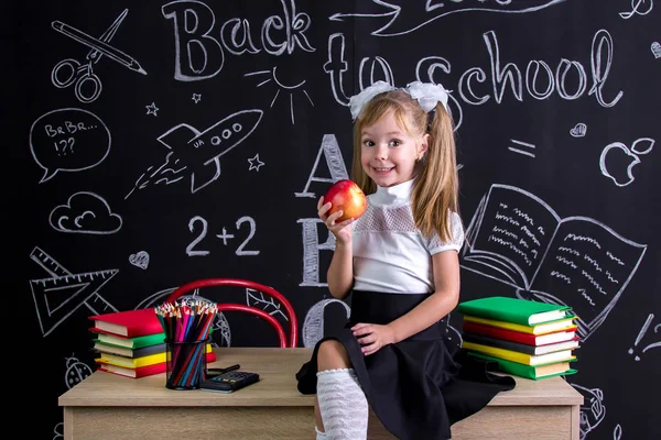 Studentessa allegra seduta sulla scrivania con libri, materiale scolastico, con una mela rossa nella mano destra — Foto Stock