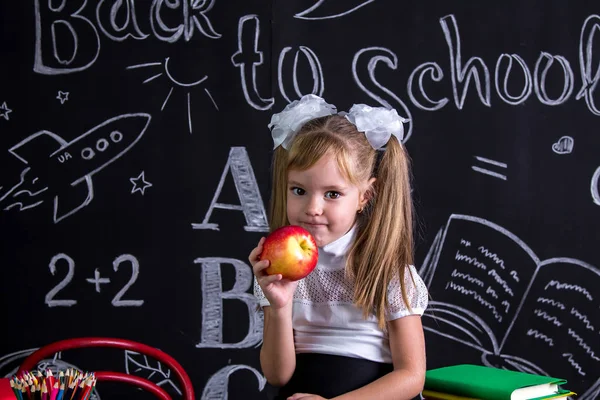 Greedy schoolgirl sitting on the desk with books, school supplies, yholding a red apple in the right hand — Stock Photo, Image
