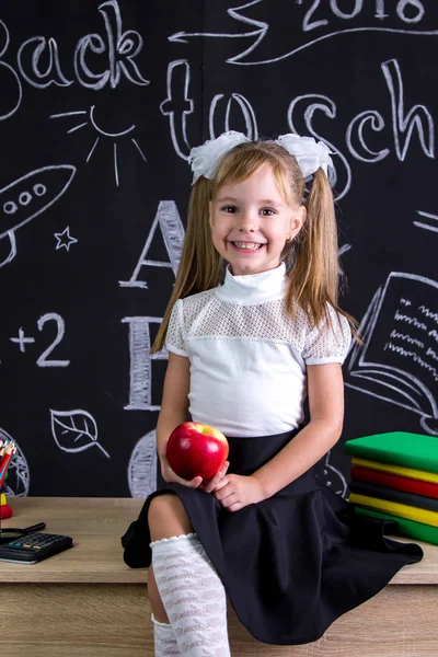 Studentessa allegra e sorridente seduta sulla scrivania con libri, materiale scolastico, con in mano una mela rossa — Foto Stock