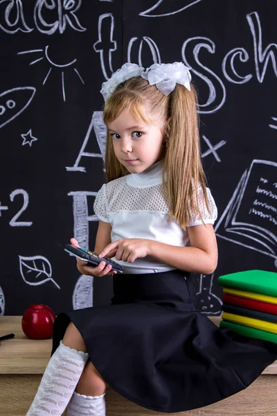 Puzzled schoolgirl sitting on the desk with books, school supplies, doing maths with calculator looking at the camera