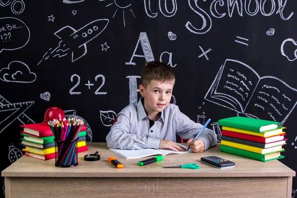 Leworęczny uczniak siedzi przy biurku z książek, przyborów szkolnych, pisząc w książce ćwiczenia, patrząc prosto do aparatu — Zdjęcie stockowe