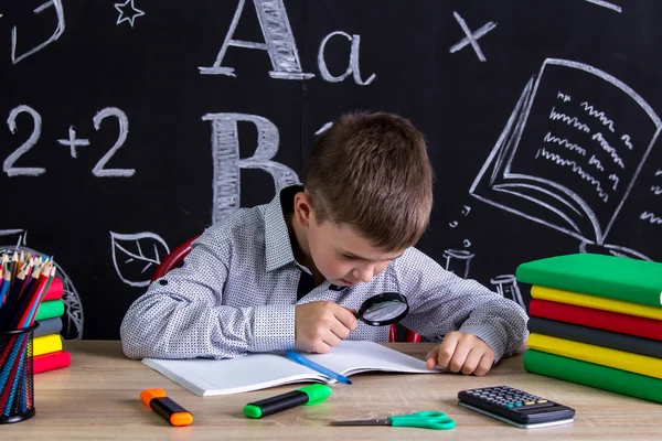 Schooljongen zitten aan de balie met boeken, schoolbenodigdheden, zoeken de tekst in het werkboek door middel van Vergrootglas. Schoolbord met schilderijen als achtergrond — Stockfoto