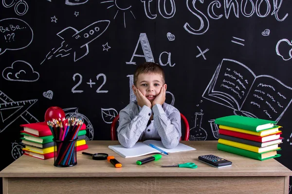 Alegre colegial sentado en el escritorio con libros, útiles escolares, con ambos brazos bajo las mejillas — Foto de Stock