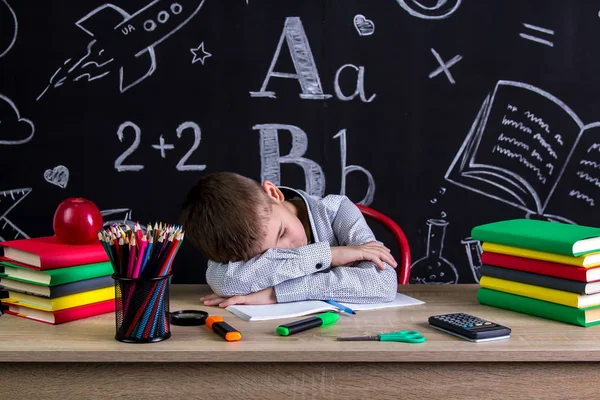 Slapen aan de balie met een hoofd schooljongen leunde op de handen, omringd met schoolbenodigdheden. Schoolbord als achtergrond — Stockfoto