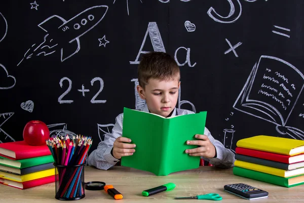 Excelente colegial sentado en el escritorio leyendo el libro, rodeado de útiles escolares. Pizarra como fondo — Foto de Stock