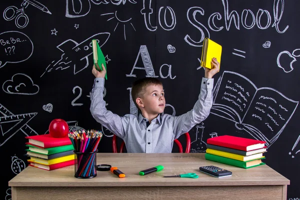 Colegial sentado en el escritorio con libros, útiles escolares, sosteniendo dos libros en sus dos brazos, mirándolos — Foto de Stock