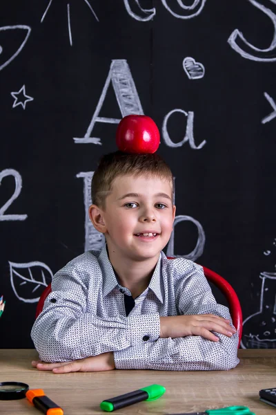 Wesoły i szczęśliwy uczniak siedzi przy biurku z książek, przyborów szkolnych, z czerwonym jabłkiem na czubku głowy. Powiększony widok — Zdjęcie stockowe