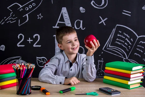 Głodny uczniak siedzi przy biurku z książki, przybory szkolne, trzymając w jej ramię Czerwone jabłko gotów gryźć — Zdjęcie stockowe