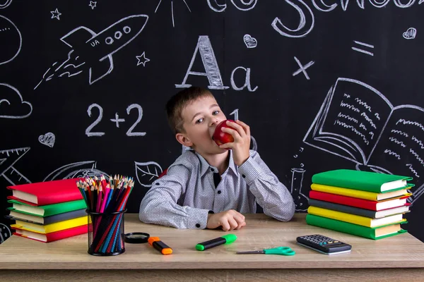 Głodny uczniak siedzi przy biurku z książki, przybory szkolne, gryzienie Czerwone jabłko, trzymając w ręku — Zdjęcie stockowe