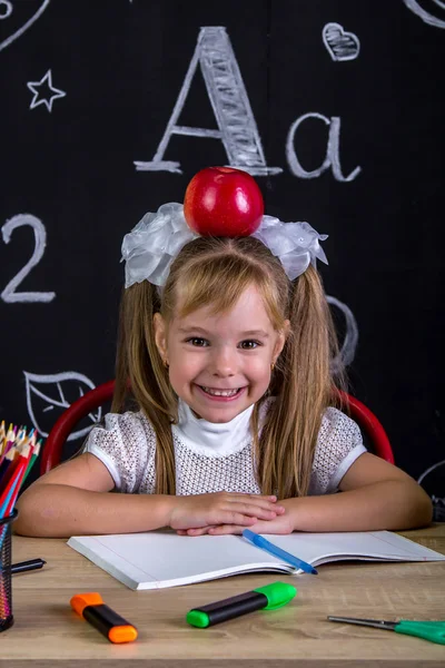 Alegre y feliz colegiala infernal sentada en el escritorio con libros, útiles escolares, con una manzana roja en la parte superior de su cabeza —  Fotos de Stock