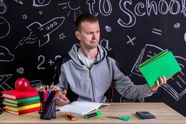 Minachting jongere zitten aan de balie met een stapel boeken in de hand, omringd met schoolbenodigdheden. Schoolbord als achtergrond — Stockfoto