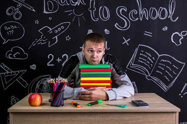 Zdziwiony chłopak siedzi przy biurku stos książek pod brodą, otoczony przyborów szkolnych. Tablica w tle — Zdjęcie stockowe