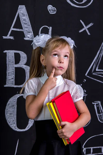 Zdziwiony uczennica stojąc przed chalkboard jako tło z kilka książek, trzymając je i wskazując palcem policzek. Obraz portret — Zdjęcie stockowe