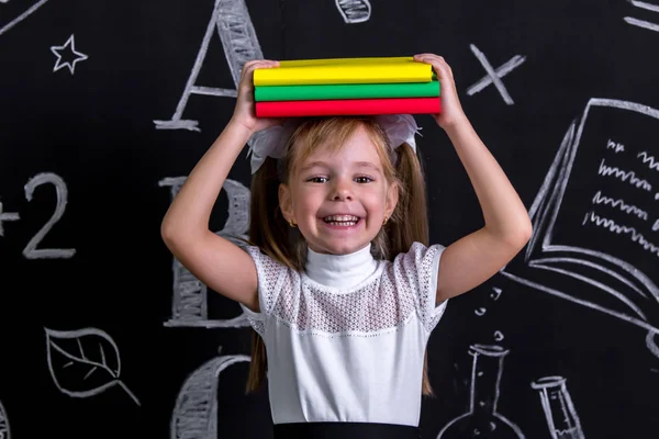 Uśmiechający się stojący szczęśliwy uczennica przed chalkboard jako tło z stos książek, trzymając je na górze głowy. Obraz portret — Zdjęcie stockowe