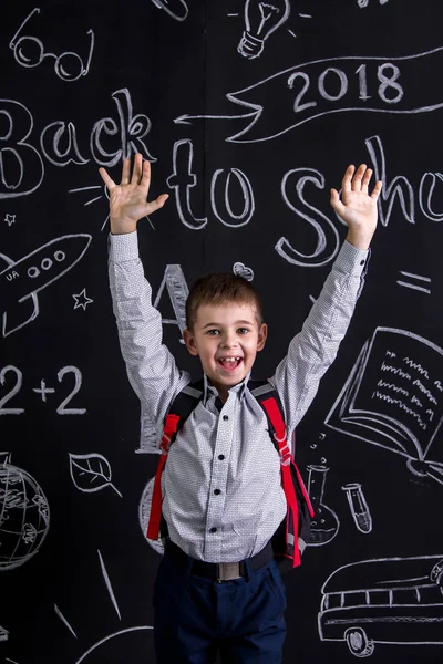 Colegial feliz y alegre de pie ante la pizarra como fondo con una mochila en la espalda y las manos levantadas. Retrato de imagen — Foto de Stock