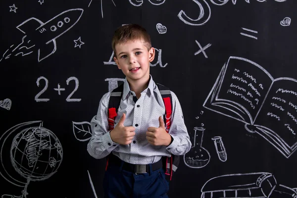 Uczniak uśmiechający się stojąc przed chalkboard jako tło z plecakiem na plecach z kciuki podniósł. Szczęście, znowu szkoła. Obraz pejzaż — Zdjęcie stockowe