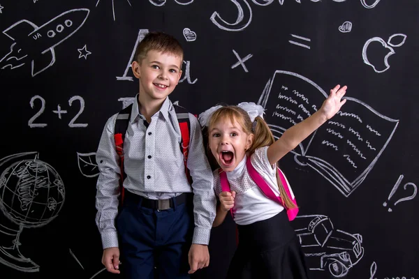 Podekscytowany i wesoły schoolkids stojąc przed chalkboard jako tło z plecakiem na plecach Wyświetlono szczęśliwy emosions. Obraz pejzaż — Zdjęcie stockowe