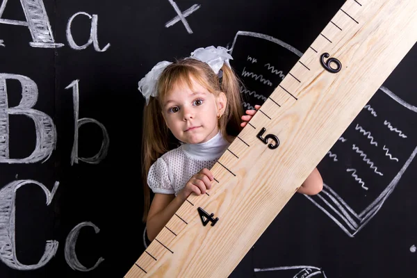 Schattig schoolmeisje staande voor het schoolbord als achtergrond de enorme liniaal schuin te houden — Stockfoto