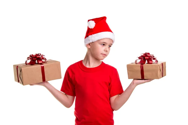 Όμορφος σοβαρές αγόρι, santa καπέλο στο κεφάλι του, με δύο κουτιά δώρων στα χέρια, ψάχνει στο αριστερό πλαίσιο. Έννοια: ευτυχισμένο το νέο έτος διακοπές τα Χριστούγεννα ή — Φωτογραφία Αρχείου
