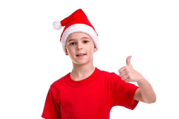 Lindo chico sonriente, sombrero de santa en su cabeza, levantando el pulgar izquierdo hacia arriba. Concepto: Navidad o Feliz Año Nuevo — Foto de Stock