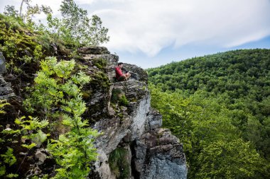 Kırmızı kazaklı ve bej pantolonlu genç bir adam yüksek bir uçurumun tepesinde oturuyor ve bir e-kitap okuyor. Kalın yeşil orman aşağılara doğru yayılmış. Cliff yosun ve küçük ağaçlarla kaplıydı. Sessizlik ve sakinlik.
