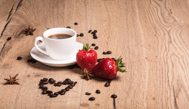 Tahta masanın üzerinde beyaz bir fincan kahve duruyor. Önlerinde kalp şeklinde serilmiş kahve çekirdekleri, anason yıldızları, iki kırmızı olgun çilek, havadar çikolata.