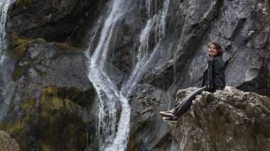Kız bir şelalenin yanındaki kayanın üzerinde oturuyor. Powerscourt Şelalesi, Wicklow Dağları, İrlanda.