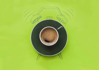 Bir fincan kahve yeşil arka planda çizilmiş bir çalar saatin üzerinde duruyor.