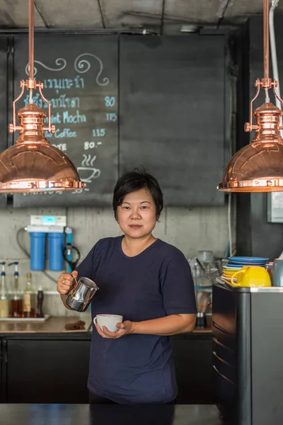 Asian woman 40s white skin work as a barista pouring latte froth to make a coffee latte art into the white coffee cup for serve to customers in the coffee shop cafe