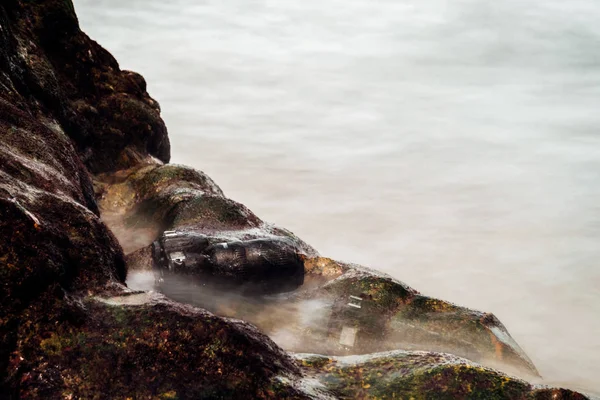 Cámara réflex digital en la playa de piedra mojada de agua mar ola —  Fotos de Stock