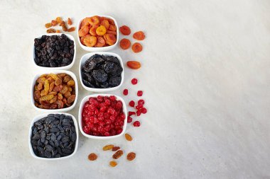 Farklı kurumuş meyveler: kuru erik, kuru üzüm, hurma, kuru kayısı ve kirazlar açık arkaplanda bir kasede, üst görünümde.