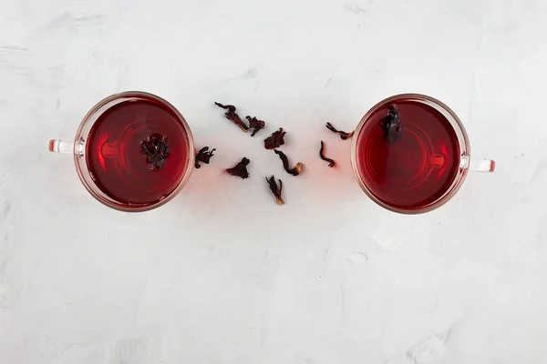 Two glass cups with red  hot hibiscus tea or sudanese rose on light grey background with dry tea leaves. Top view.