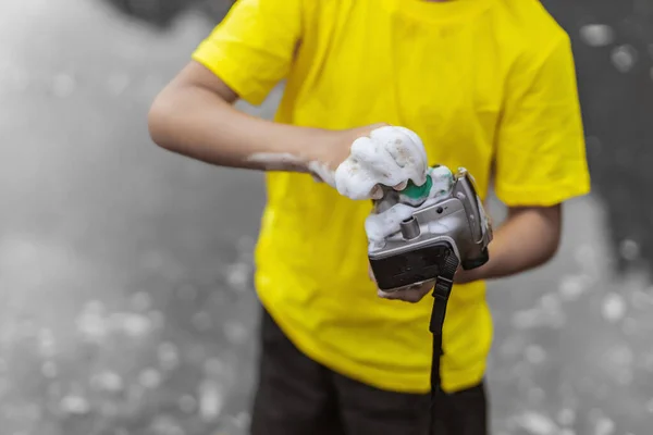 A schoolboy in a yellow t-shirt and shorts is standing near the water in nature and washing a camera with soap and foam. The concept joke and humor. Bright color photo. Close up.
