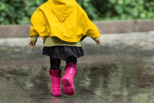 Sarı yağmurluklu ve pembe lastik çizmeli mutlu küçük bir kız sokakta tek başına yağmurda yürüyor. Park, doğa. Çocukluk konsepti. Evrensel Çocuk Günü.