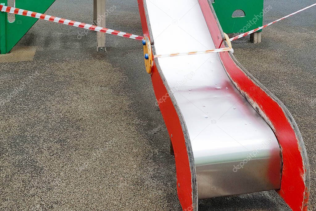 Red metal slide for children to ride on the playground in the park wrapped with red barrier tape. Outside. Prohibition of outdoor walks, prevention of the coronavirus influenza virus covid-19.