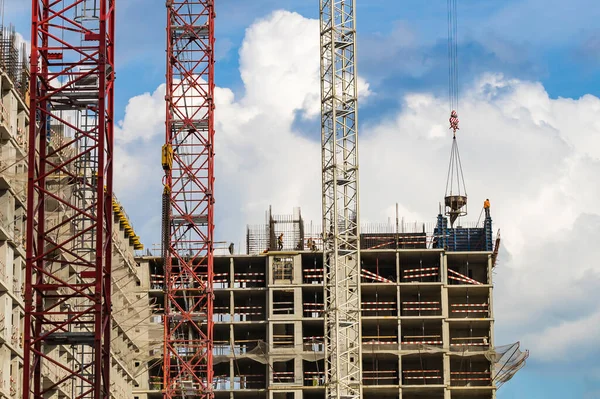 Several red cranes have been put to work on the construction of a modern multi-storey residential complex against a blue sky with white clouds. Concept work for builder. Outdoors. Summer day.