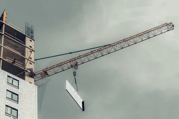 One high-rise crane hauling glare plate against a house and gray sky during the construction phase. Industry concept for low-income young families. Mortgage, business, real estate loan. Copy space.