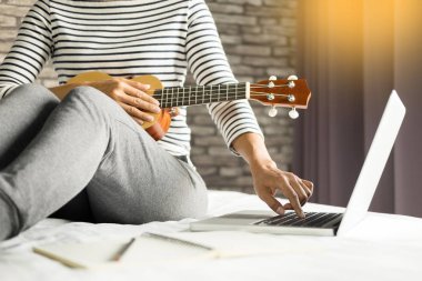 Mutlu Genç Asyalı kadın yatakta yatak odasında oturmuş ukulele oynarken