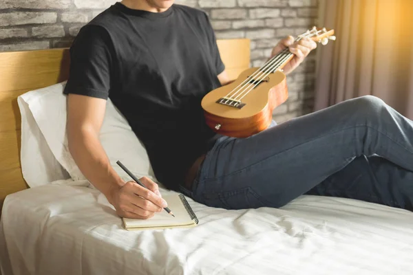 幸せな若いアジア男の寝室のベッドの上に座ってウクレレを演奏 — ストック写真
