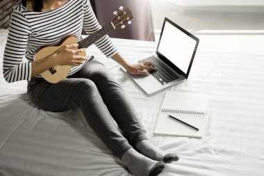 Mutlu Genç Asyalı kadın yatakta yatak odasında oturmuş ukulele oynarken