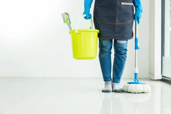 Young housekeeper cleaning floor mobbing holding mop and plastic bucket with brushes, gloves and detergents in the leaving room house floor helping his wife