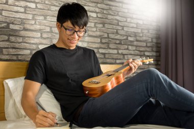 Mutlu genç Asyalı adam yatakta yatak odasında oturmuş ukulele oynarken