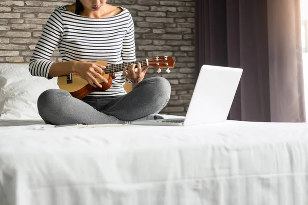 Happy young asian woman playing ukulele sitting on bed in bedroo — Stock Photo, Image