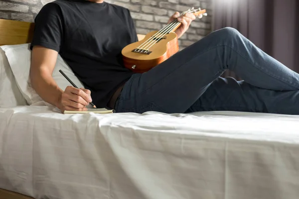 Šťastný mladý Asiat, hraje na ukulele, sedí na posteli v ložnici. — Stock fotografie