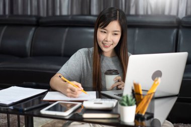 Attractive young beautiful asian woman working with laptop and document drinking coffee at the indoors living room office as a freelancer, e-coaching working, remotely or work from home concept clipart