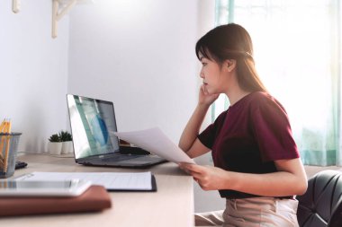 Çekici, genç, güzel Asyalı kadın ev içi oturma odasında bağımsız olarak otururken, uzaktan ya da ev kavramından bağımsız olarak çalışırken dizüstü bilgisayar ve belgeyle çalışıyor.