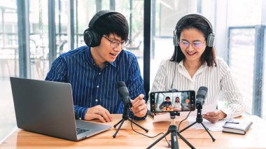 Asyalı erkek ve kadın podcastları kulaklıklarda iş arkadaşlarıyla birlikte mikrofon ve kameraya konuşma, iletişim teknolojisi ve eğlence konseptini kaydediyorlar.